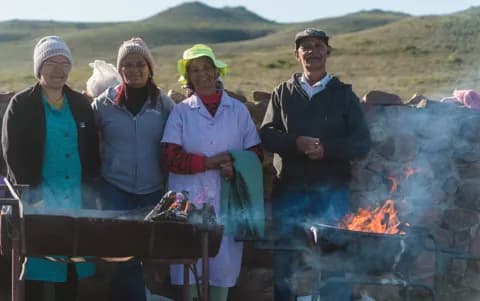 Four people cooking roosterkoek over an open fire in front of green hills