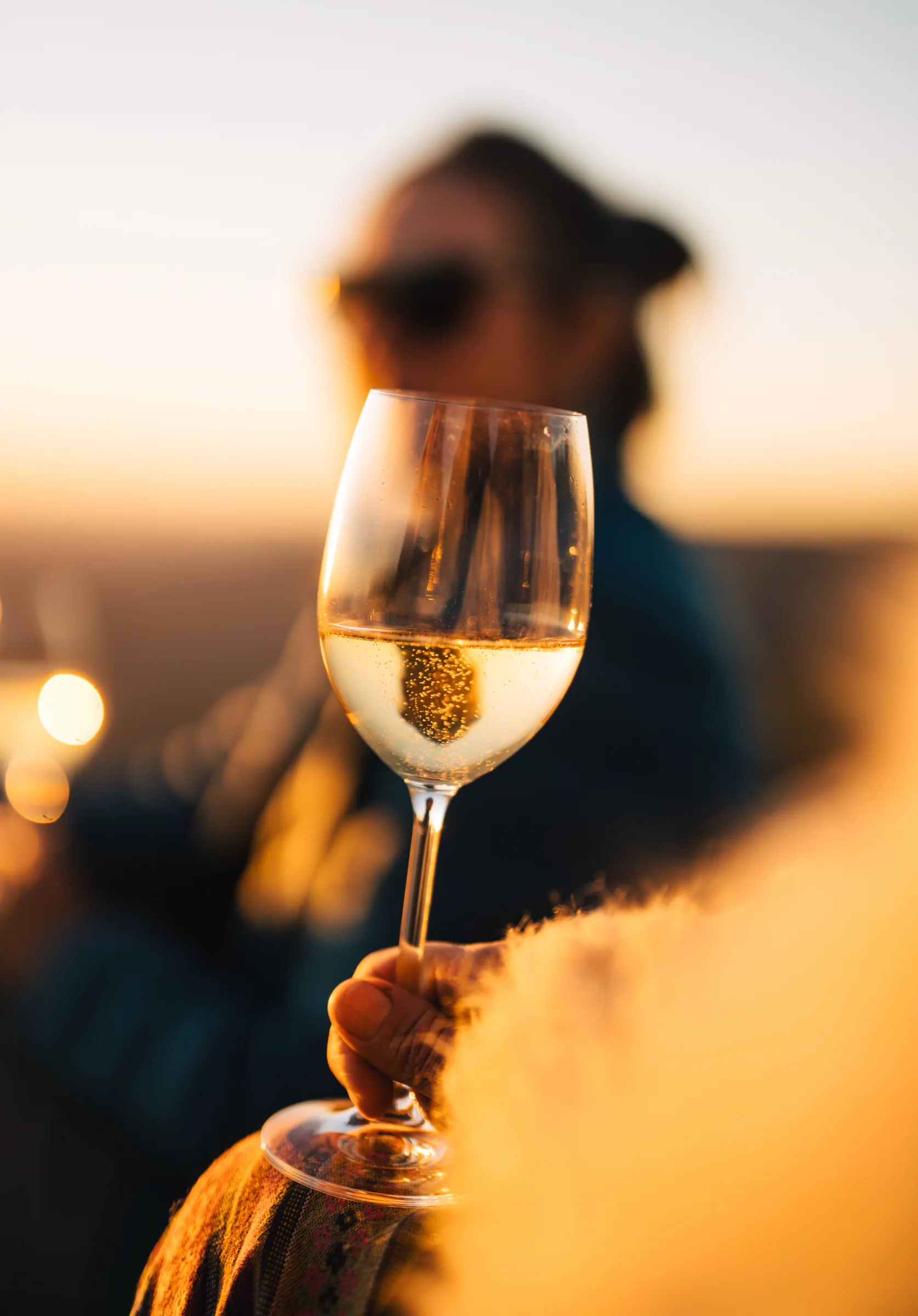 A glass of white wine held up in evening light, a person out of focus behind
