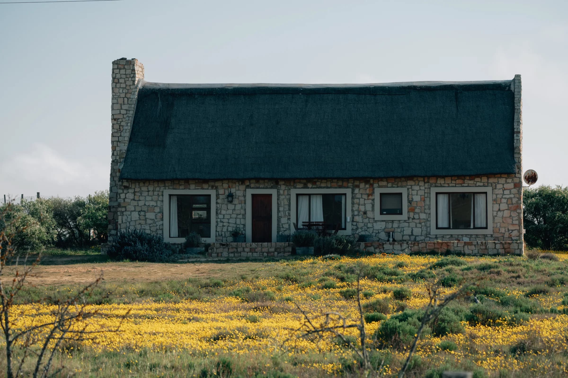 Kliphuis, the Stone Cottage: a thatched stone house with a chimney, in a field of yellow flowers